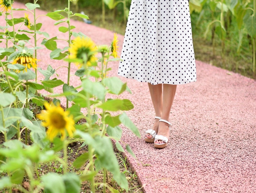 Nahaufnahme einer Person in weißem Kleid mit schwarzen Punkten und weißen Sandalen, die auf einem rosafarbenen Kiesweg zwischen blühenden Sonnenblumen steht.